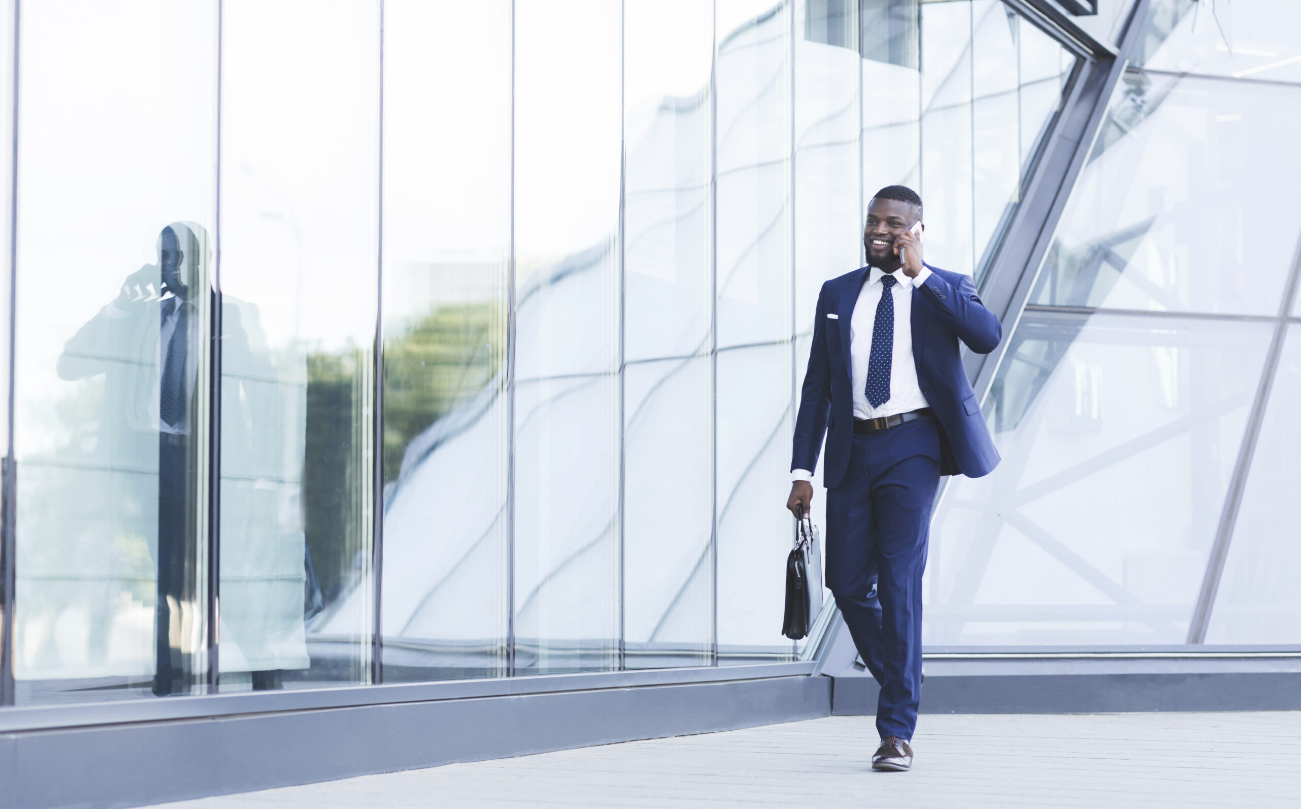 A young business professional walking outside an office, talking on his mobile phone while holding a briefcase.