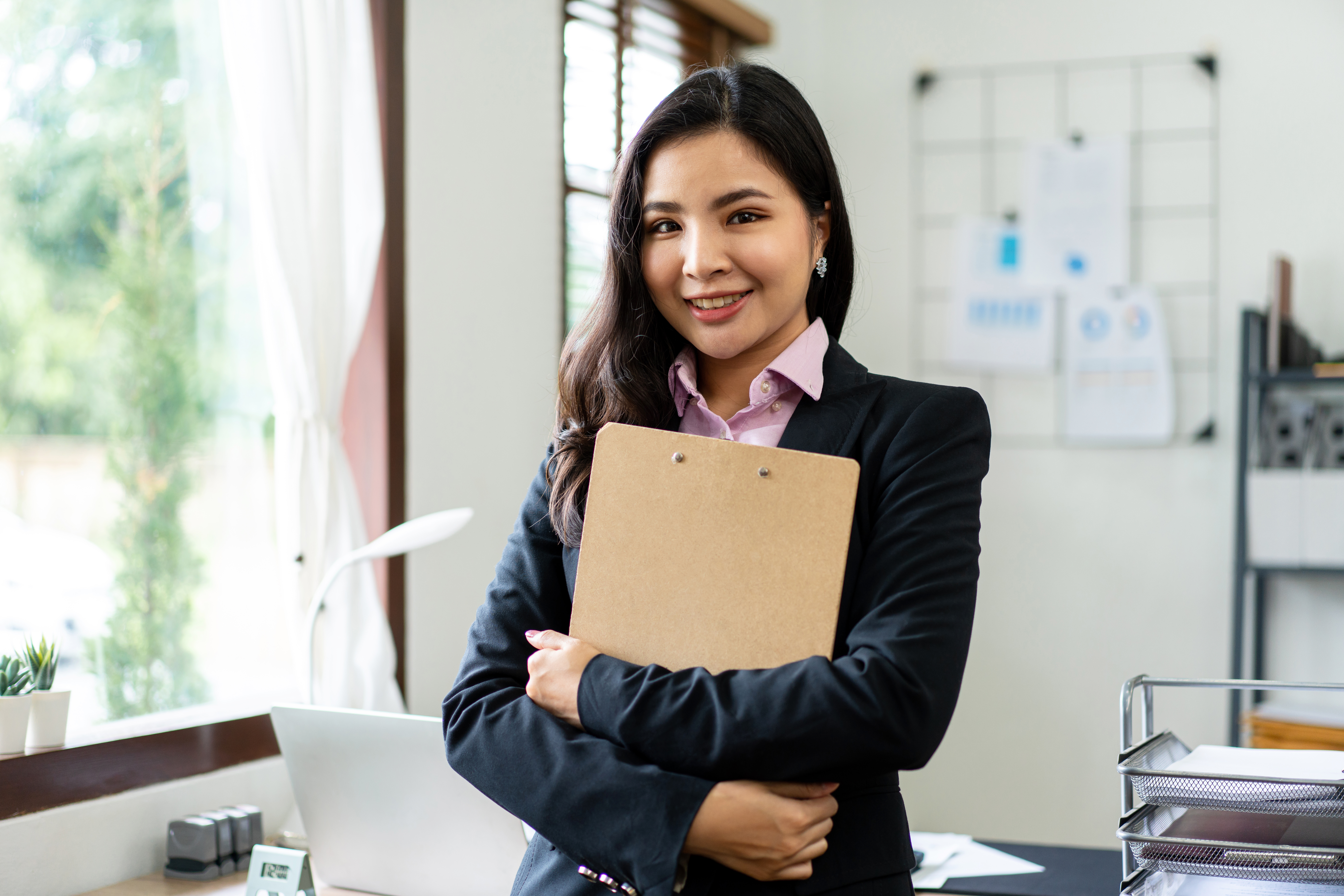 A smiling young professional standing in an office setting and holding a clipboard