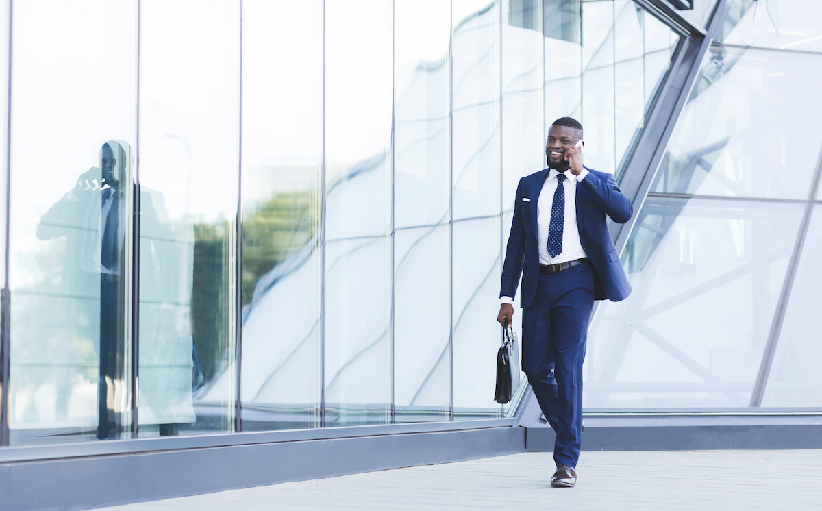 A young business professional walking outside an office, talking on his mobile phone while holding a briefcase.